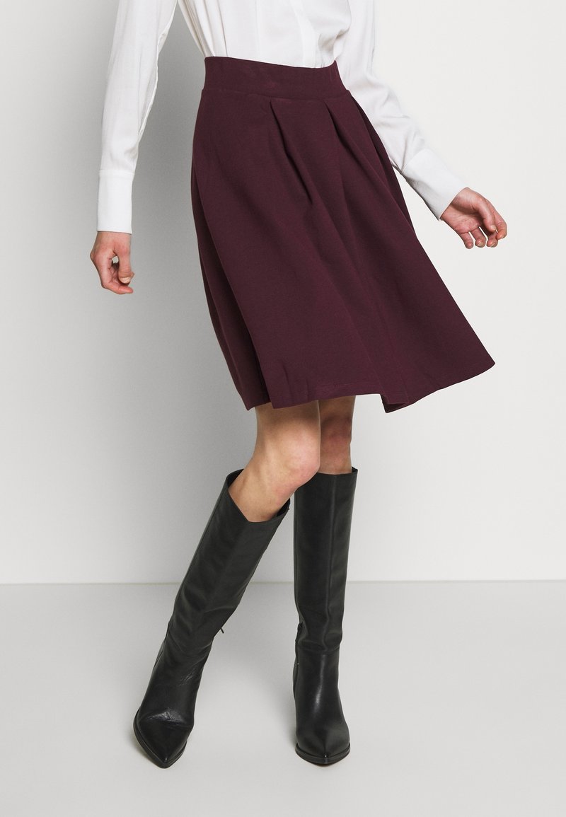 Femme portant une jupe plissée bordeaux longueur genou, une blouse blanche à manches longues et des bottes noires montantes jusqu’aux genoux, debout devant un fond uni.