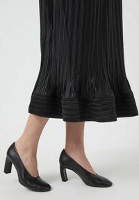 Sorte læder sko med høje hæle, en buet halsudskæring og en firkantet hæl, kombineret med et plisseret sort skørt med horisontal tekstur.