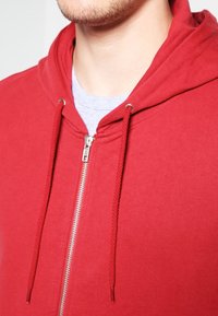 Sudadera roja con cremallera y capucha estructurada, con cordones y detalles de cremallera plateada. Hecha de una tela suave con una textura lisa.