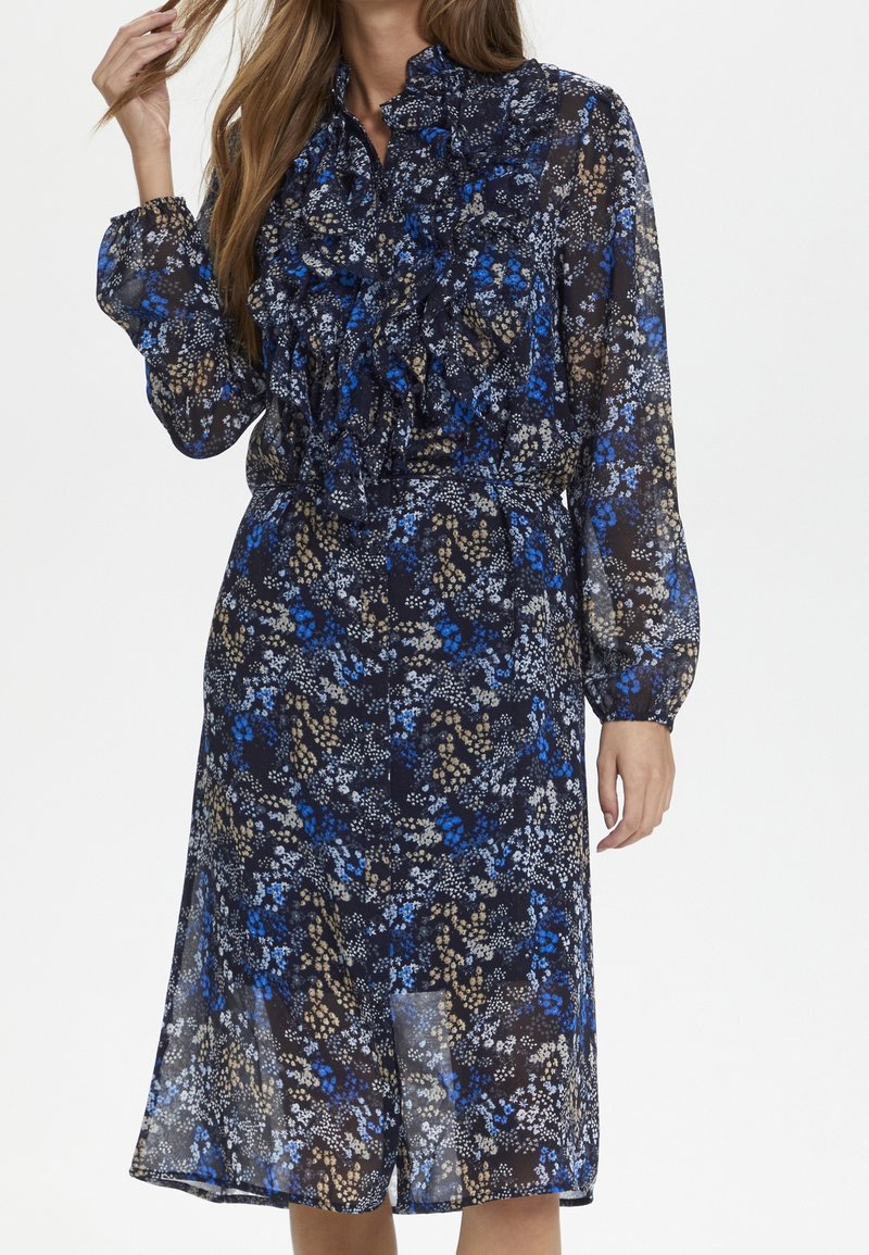 Robe fleurie à manches longues et longueur genoux avec des motifs bleus, blancs et beiges, dotée d'un devant à volants et d'une taille cintrée.