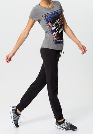 Frau, die ein graues Grafik-T-Shirt, schwarze Jogginghosen und schwarz-weiße Sneakers trägt, vor einem schlichten weißen Hintergrund.