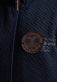 Marinblå quiltad jacka med en rund brun patch med en örn och rutmönster, kompletterad med en blå dragkedja och präglad text.