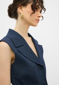 Marinblå denim utan ärm blazer med breda slag och djup V-ringning. Har en enda knappstängning och en strukturerad tygfinish. Guld-ringarörhängen.