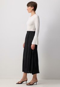 Vit långärmad topp med veckade manschetter, parat med en svart veckad midikjol. Bärs med svarta klacksandaler, vilket visar en minimalistisk estetik.