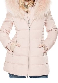 Mujer vestida con un abrigo de invierno acolchado color rosa claro con capucha de piel, con las manos en los bolsillos y jeans azules visibles por debajo.