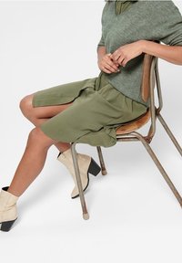 Nainen istuu ruosteisessa metallituolissa, päällä vihreä mekko, harmaa neuletakki ja vaaleanbeige nilkkasaappaat, joissa on paksut korot.