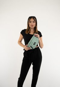 Borsa con motivo geometrico verde e etichetta nera, tenuta davanti a uno sfondo uniforme; presenta un design elegante e curvo con cerniera.