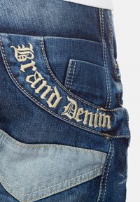 Jeans in denim con tessuto blu scuro, accenti di blu chiaro in contrasto e testo ricamato "Grand Denim" sulla tasca posteriore.