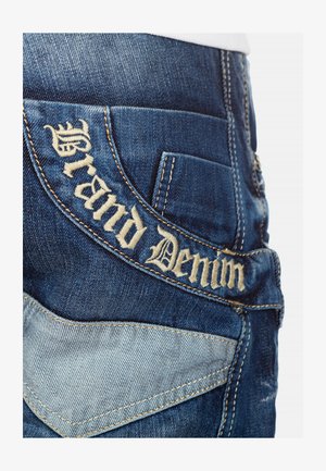 Denim jeans met donkerblauwe stof, contrasterende lichtblauwe accenten en geborduurde "Grand Denim" tekst op de achterzak.