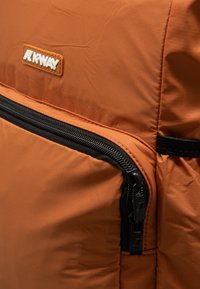 Zaino K-Way arancione con tessuto testurizzato, dettaglio della cerniera prominente in nero e un logo in patch bianco applicato sul davanti.