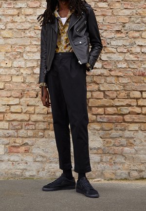 Svart lädermotorcykeljacka med silverdetaljer, blommig skjorta med knappar, svarta byxor och svarta sneakers på en texturerad tegelvägg.