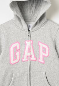 Grå zip-opp hettegenser med en rosa "GAP" logo på forsiden, med hette og lommer, laget av mykt stoff med en glatt tekstur.
