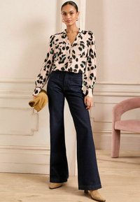 Blusa rosa a maniche lunghe con stampa animale nera e verde, abbinata a jeans a vita alta in denim scuro a zampa d'elefante. Tiene una clutch beige.