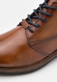 Zapato de cuero marrón con una textura suave, con cordones azul oscuro y un diseño cosido. La puntera es redondeada y la suela es negra.