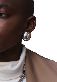 Orecchino grande e rotondo in argento con una superficie lucida che riflette l'ambiente circostante. Indossato da una persona con un taglio di capelli corto, abbinato a un abbigliamento neutro.