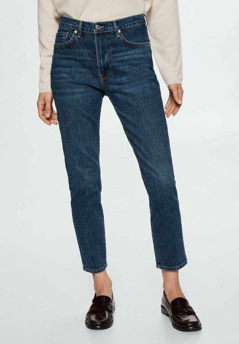 Adulte portant un jean droit bleu foncé, un haut beige à manches longues et des mocassins en cuir marron foncé, debout devant un fond uni.