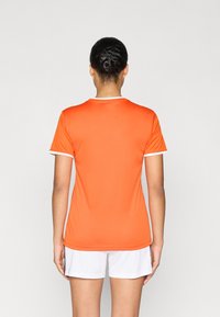 Πορτοκαλί αθλητική φανέλα από λείο ύφασμα, με κοντά μανίκια, στρογγυλή λαιμόκοψη και λευκή λεπτομέρεια στο γιακά και τα μανίκια.