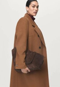 Bolso de ante marrón sostenido por una persona que lleva un abrigo color camello. El bolso cuenta con una solapa plegable y tiene una superficie suave y texturizada.