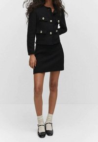 Mujer vestida con un traje de falda texturizada negra con botones dorados, combinado con calcetines blancos y zapatos Mary Jane negros, de pie contra un fondo liso.