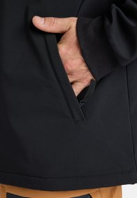 Black softshell jacket with a side pocket. Features include a smooth texture, ribbed cuff, and contrasting tan at the hem.