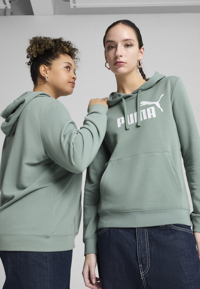 Felpe con cappuccio verde chiaro, dotate di tasche frontali e con il logo PUMA bianco. Un modello indossa una gonna di jeans; l'altro ha i capelli ricci.