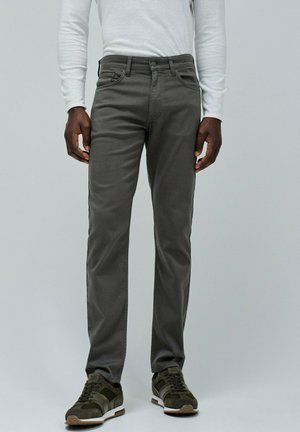 Hombre vestido con pantalones ajustados gris oscuro, camisa de manga larga blanca y zapatillas verde oscuro, de pie contra un fondo claro y liso.
