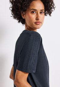Tmavě modrý top s krátkým rukávem a texturovanými akcenty na ramenou, kulatým výstřihem a jemným materiálem. Jednoduchá, elegantní silueta s minimálními detaily.