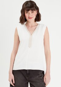 Top blanc sans manches avec un décolleté en V, exhibant un détail de bord texturé au centre. Tissu doux avec une coupe ample.