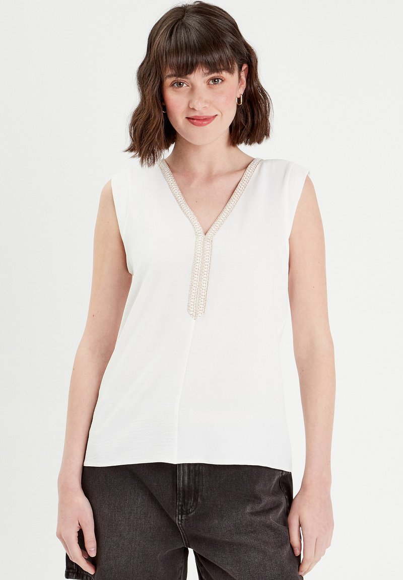 Top blanc sans manches avec un décolleté en V, exhibant un détail de bord texturé au centre. Tissu doux avec une coupe ample.
