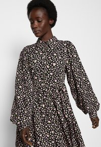 Vestido preto floral com decote tipo colarinho, mangas longas bufantes e cintura franzida. O padrão inclui pequenas flores rosa e verdes.