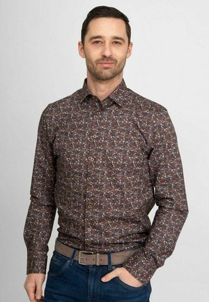 Man met kort donker haar draagt een bruin patroonoverhemd met lange mouwen, in een blauwe spijkerbroek gestopt met een grijze riem, hand in zak.