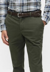 Man i gröna byxor med ett brunt flätat läderbälte och en långärmad rutig skjorta, händerna delvis i fickorna.