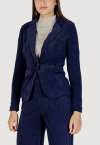 Námořnický kordurový blazer s přiléhavým střihem, klopami a předními kapsami, spárovaný se světle zbarveným topem s vysokým límcem.