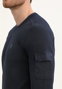 Temno modra majica z dolgimi rokavi iz teksturiranega materiala, s krožnim izrezom in žepom na rokavu, ki ima dva gumbka.