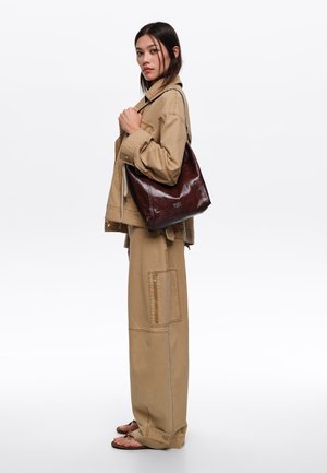 Mujer con chaqueta y pantalones utilitarios color camel, sosteniendo un bolso de cuero marrón oscuro al hombro, de pie frente a un fondo blanco liso.