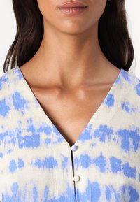 Blå og beige tie-dye topp med V-halsdesign, med to knapper ved halsen og teksturert stoff.