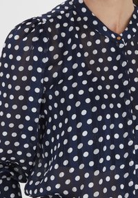 Blouse marine en tissu transparent avec des pois blancs, présentant un col rond et des manches bouffantes froncées pour une apparence texturée.