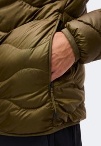 Main dans la poche zippée d'une veste matelassée vert olive avec des lignes de couture visibles et un petit logo en tissu sur le poignet.