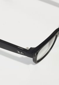 Occhiali da sole neri rettangolari con accenti in metallo, lenti trasparenti e logo Ray-Ban sulla astina, che mostrano un design elegante e una finitura opaca.