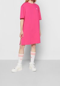 Rosa oversized t-shirt klänning med korta ärmar, sidofickor och en liten vit logotyp på bröstet, kombinerad med vita sneakers och randiga strumpor.