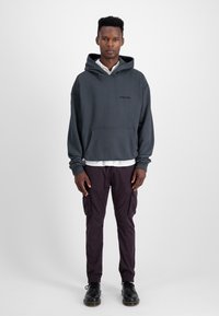 Grijze hoodie met voorzak, gecombineerd met donkerpaarse cargobroek en zwarte schoenen. Model staat tegen een effen achtergrond.