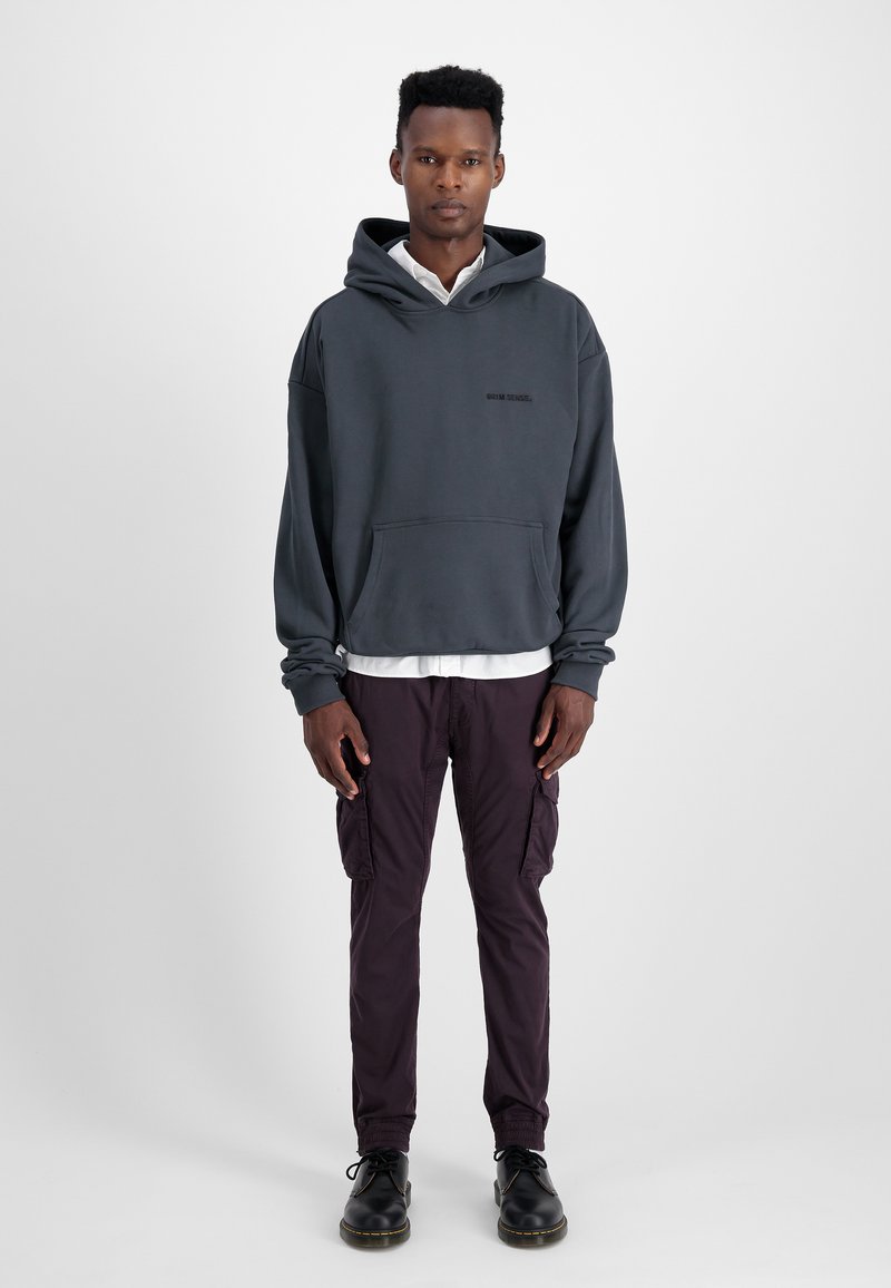 Szara bluza z kapturem, z kieszenią z przodu, w połączeniu z ciemnofioletowymi spodniami cargo i czarnymi butami. Model stoi na tle gładkiej ściany.