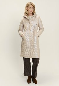 Donna che indossa un lungo cappotto trapuntato beige con cappuccio, mani nelle tasche, pantaloni a quadretti marroni e stivali alla caviglia marroni, in piedi contro uno sfondo semplice.