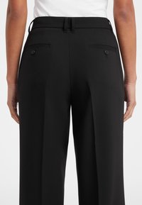 Pantaloni neri su misura con una consistenza liscia, gamba larga e due tasche posteriori con bottoni. Presentano passanti per la cintura in vita.