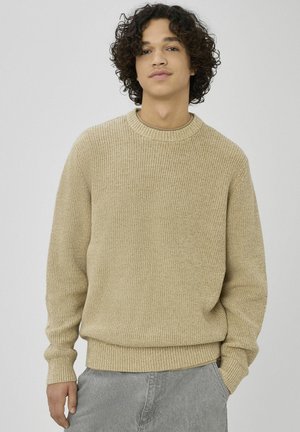 Jeune homme aux cheveux bouclés portant un pull en maille beige et un pantalon gris clair, debout avec une main dans sa poche.