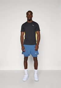 Must sportlik t-särk kollaste küljesuurendustega, siniste lühikeste pükstega. Mõlemal on Puma logo. Modell kannab valgeid sokke ja jalatseid.