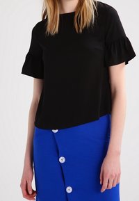 Femme portant un haut noir à manches courtes avec des volants aux manches et une jupe bleue boutonnée, debout contre un fond clair uni.