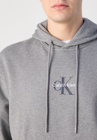 Zbliżenie na osobę noszącą szarą bluzę z kapturem z troczkami i logo Calvin Klein na piersi.