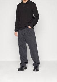 Svart långärmad pikétröja med krage och subtil logotyp, parat med löst sittande mörkgrå denimbyxor och svarta robusta skor.