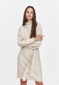 Beiges Strickkleid mit hohem Kragen, langen Ärmeln und einem Bindegürtel. Strukturierter Stoff mit lockerer Passform, der bis zur Mitte des Oberschenkels reicht.